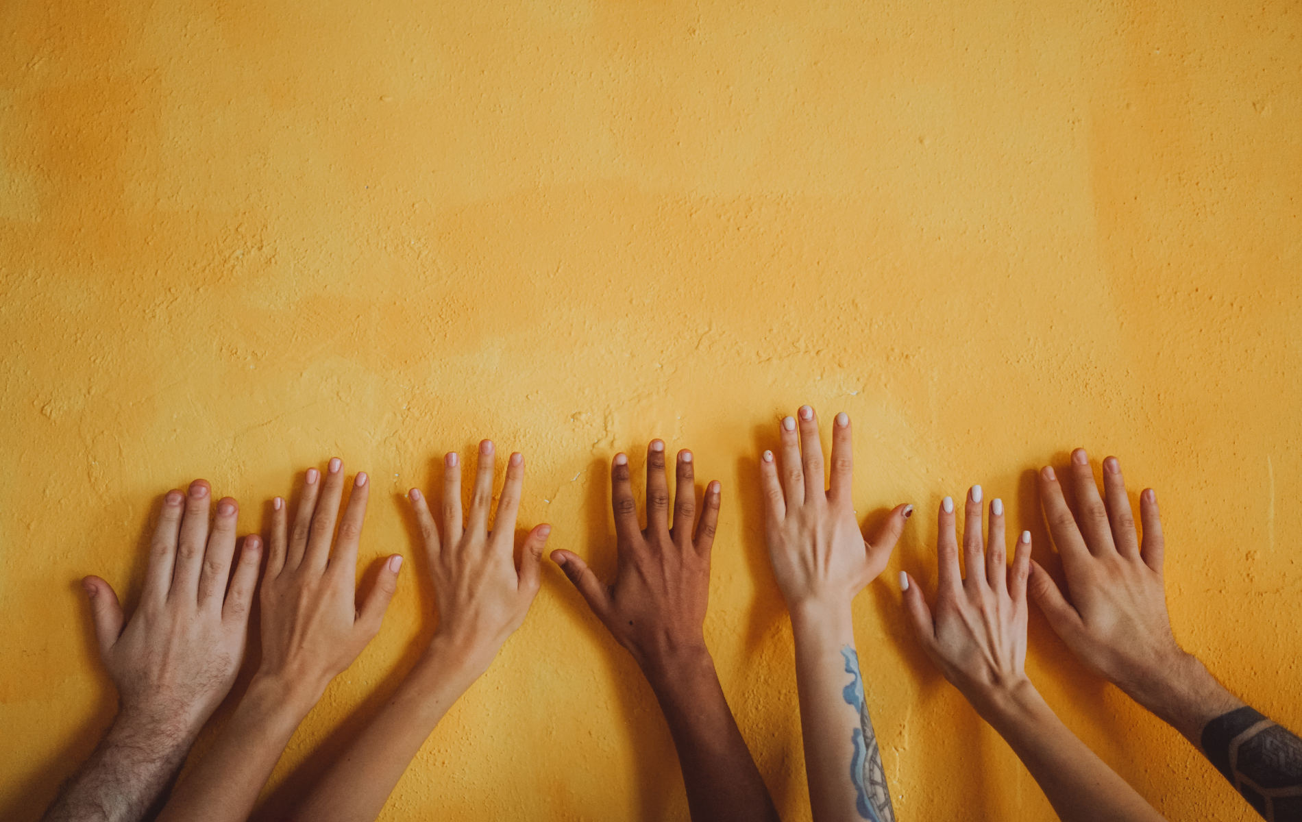 Seven hands pressed up against a bright yellow wall.