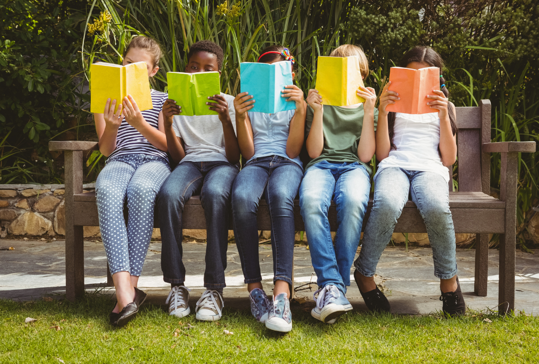A group of children sit on a bench outside reading from copy books.