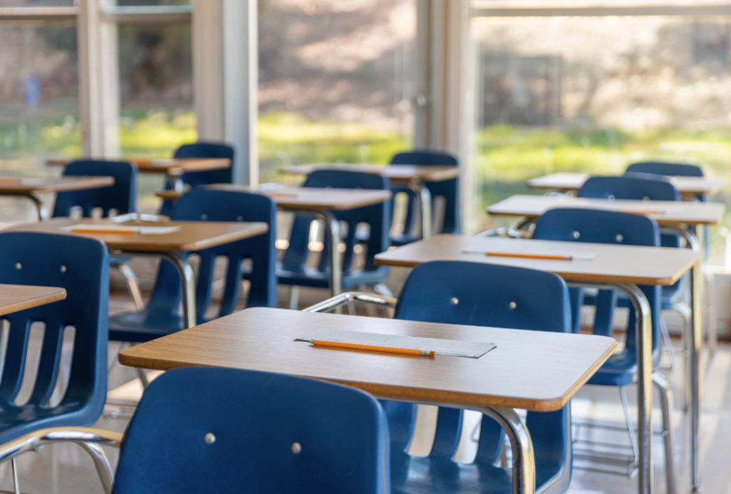 An empty classroom with tables and chairs. There is a pencil and sheet of paper on every desk
