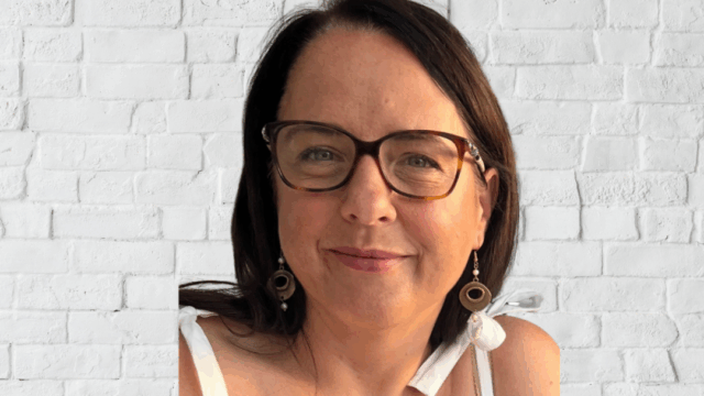 Jayne is positioned in front of a white brick wall, shown from the shoulders up. Jayne has shoulder‑length dark hair and is wearing a sleeveless white top with tied straps.