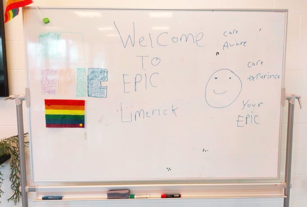 Care Community Limerick Whiteboard with “Welcome to EPIC Limerick” handwritten in the centre, surrounded by colourful drawings, a rainbow flag pinned on the left, and words like “care,” “aware,” and “experience” written around a smiley face.