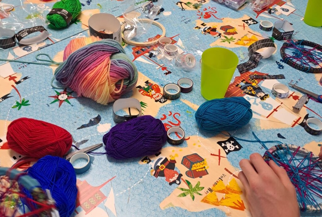 Care Community Wool Craft table covered with colourful yarn, tape, and handmade weaving projects, including circular frames being decorated by participants.