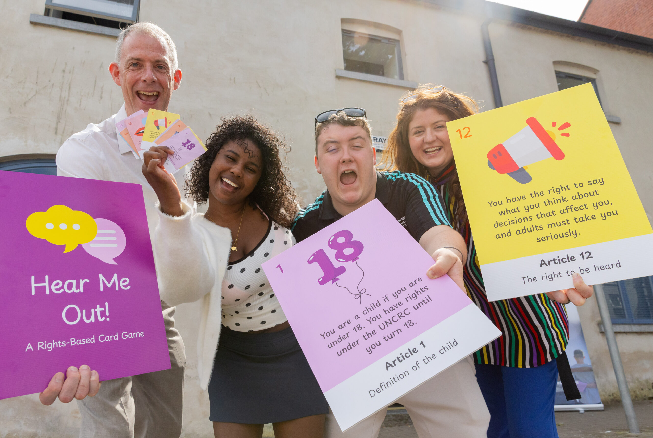 Four people pose outdoors in front of a light-colored building while holding large, colorful cards and a card game display. On the left, a purple sign reads “Hear Me Out! A Rights-Based Card Game” with speech-bubble graphics. One person holds several smaller game cards. In the center, a large pink card displays the number “18” and the text: “You are a child if you are under 18. You have rights under the UNCRC until you turn 18.” The card is labeled “Article 1 – Definition of the child.” On the right, a yellow card with a megaphone illustration states: “You have the right to say what you think about decisions that affect you, and adults must take you seriously.” The card is labeled “Article 12 – The right to be heard.” Bright sunlight creates lens flare across the image, highlighting the educational and advocacy-focused display.