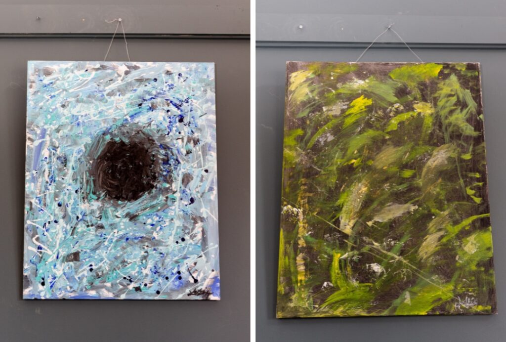 Two abstract canvases hang on a dark grey wall. The painting on the left features layers of light blue, white, black, and turquoise brushstrokes surrounding a dark circular centre. The painting on the right uses green, yellow, brown, and black tones in energetic, overlapping brushstrokes that evoke dense foliage or natural textures.