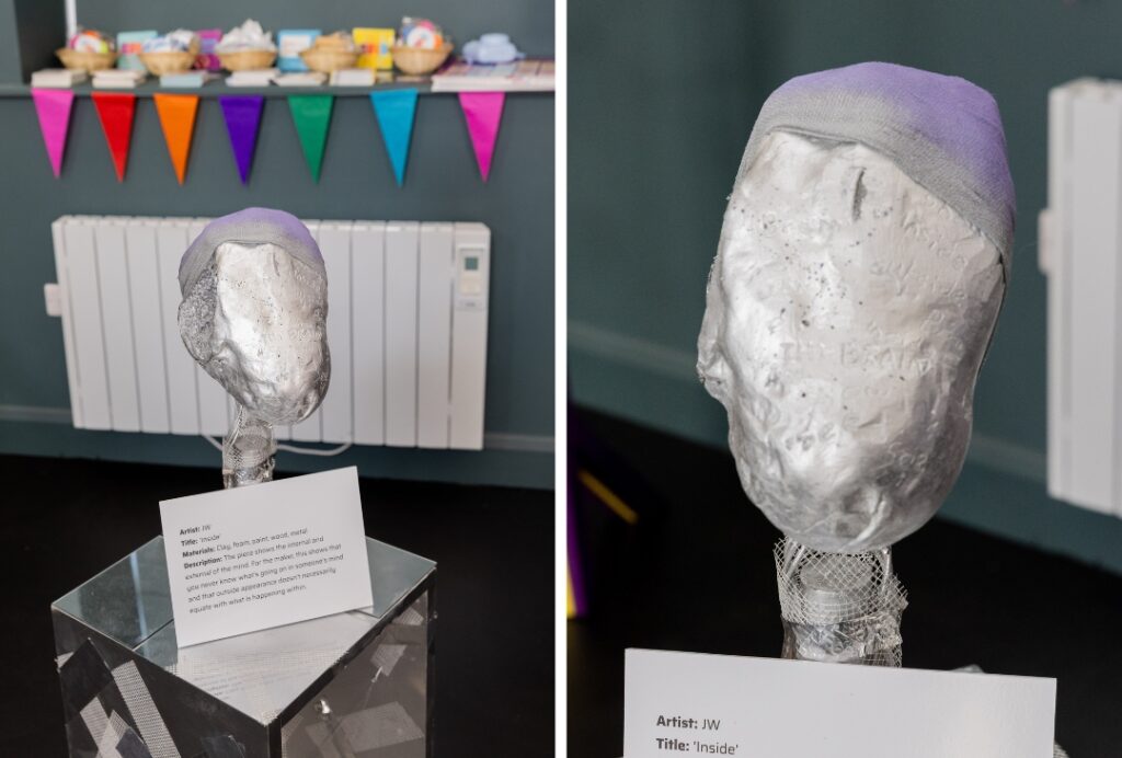 A sculpted white head mounted on a wire mesh stand is displayed on a clear acrylic pedestal. A light purple fabric covers the top portion of the head. An accompanying card identifies the artwork as “Inside” and notes that it is made from clay, foam, paint, wood, and metal. Colourful bunting and a white radiator are visible in the background. A close-up image shows the textured surface of the head sculpture in greater detail.