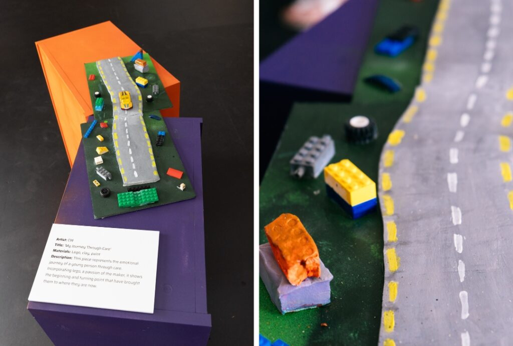 A mixed-media artwork displayed on a purple and orange pedestal features a winding grey road with white lane markings and yellow edges. Small toy building blocks, miniature vehicles, and scattered pieces line both sides of the road. A display card identifies the piece as “My Journey Through Care” and describes it as representing a young person’s emotional journey through care. A close-up view highlights the painted road and colourful block elements.