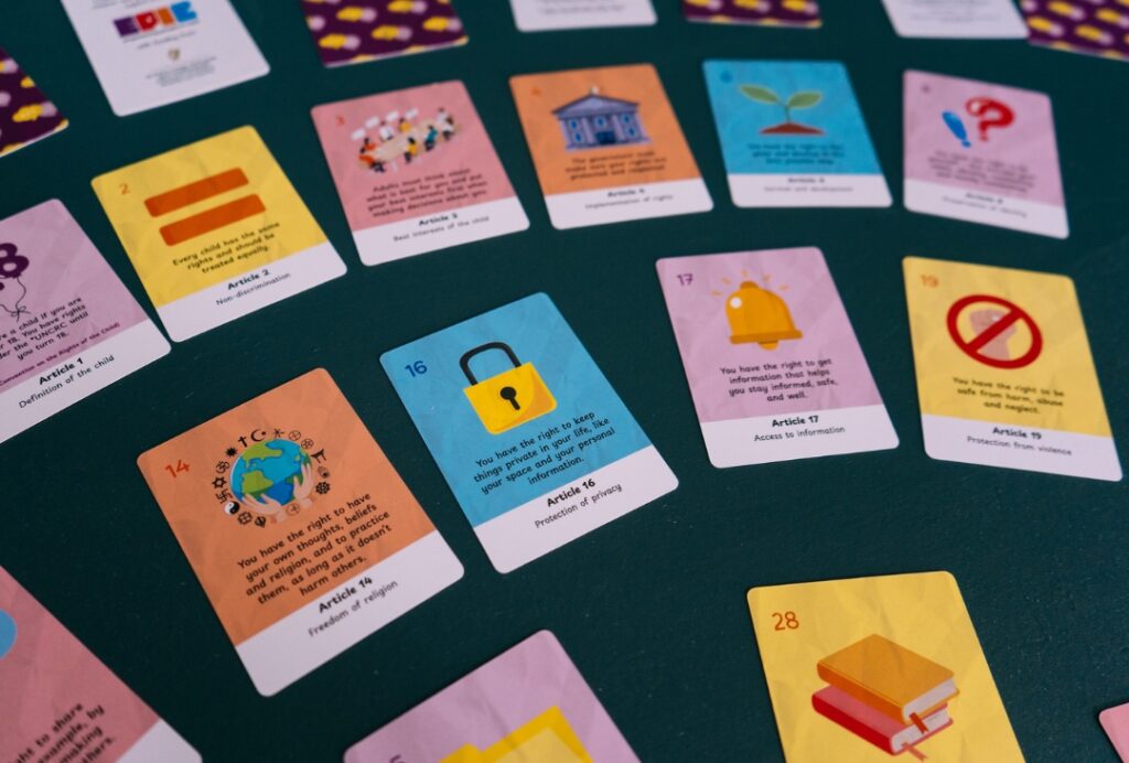 Colourful educational cards are arranged across a dark green tabletop. Each card illustrates a different children’s rights article with simple icons and short descriptions. Visible cards include Article 14 (Freedom of religion), Article 16 (Protection of privacy) with a yellow padlock icon, Article 17 (Access to information) with a bell icon, and Article 19 (Protection from violence) with a prohibition symbol. Additional cards featuring equality, participation, and education themes are scattered around the display.