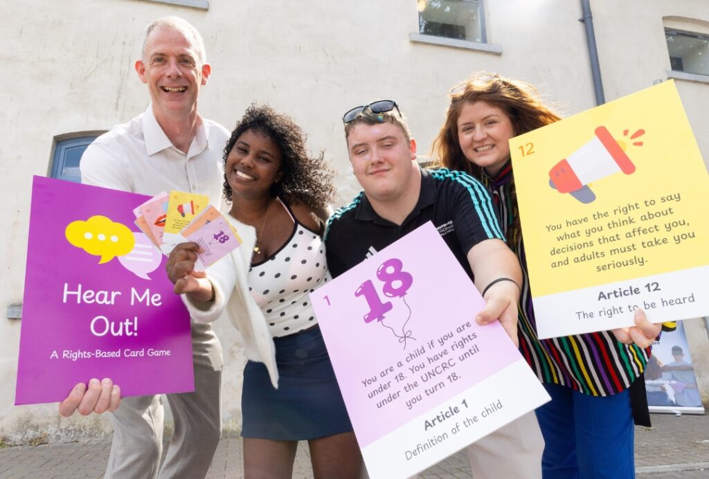 Four people outdoors holding large children's rights cards and a sign for the Hear Me Out! card game.