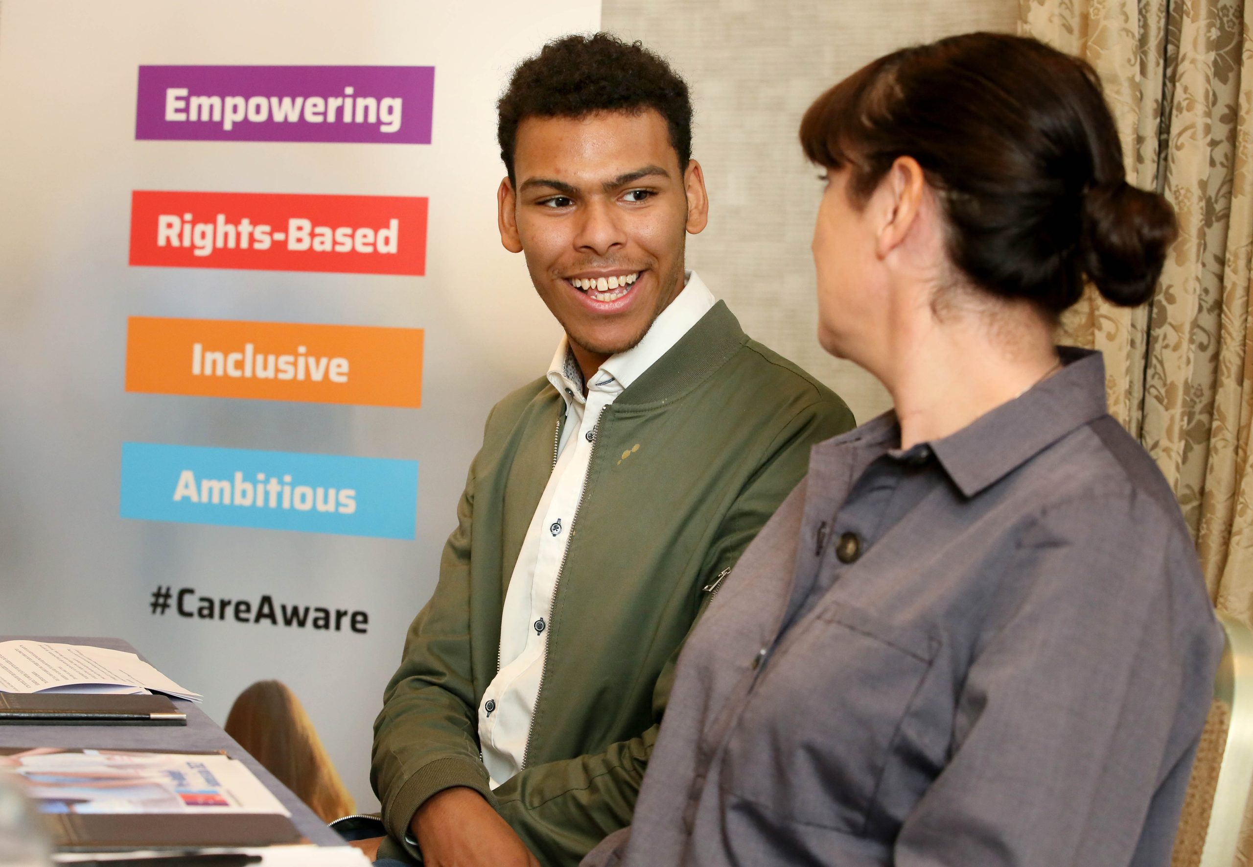 A young man wearing a white shirt and green jacket smiles while talking to a woman in a grey shirt.