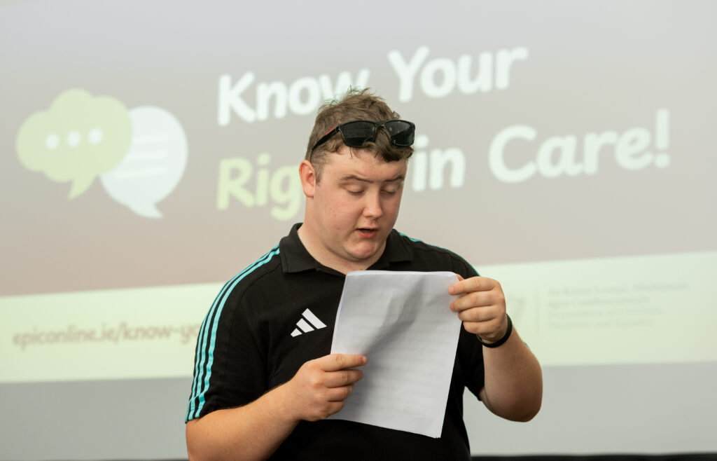 A young man reads from a speech in front of a screen that says 'Know Your Rights in Care'
